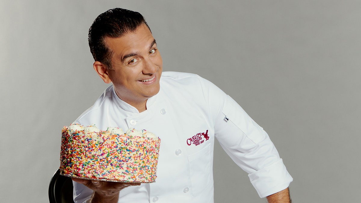Buddy Valastro soft smiles and holds a rainbow sprinkled cake