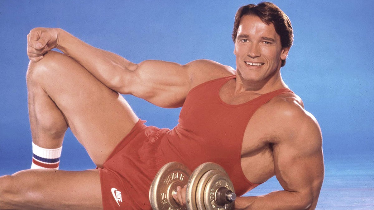 Arnold Schwarzenegger holds dumbbells while posing in red fitness gear