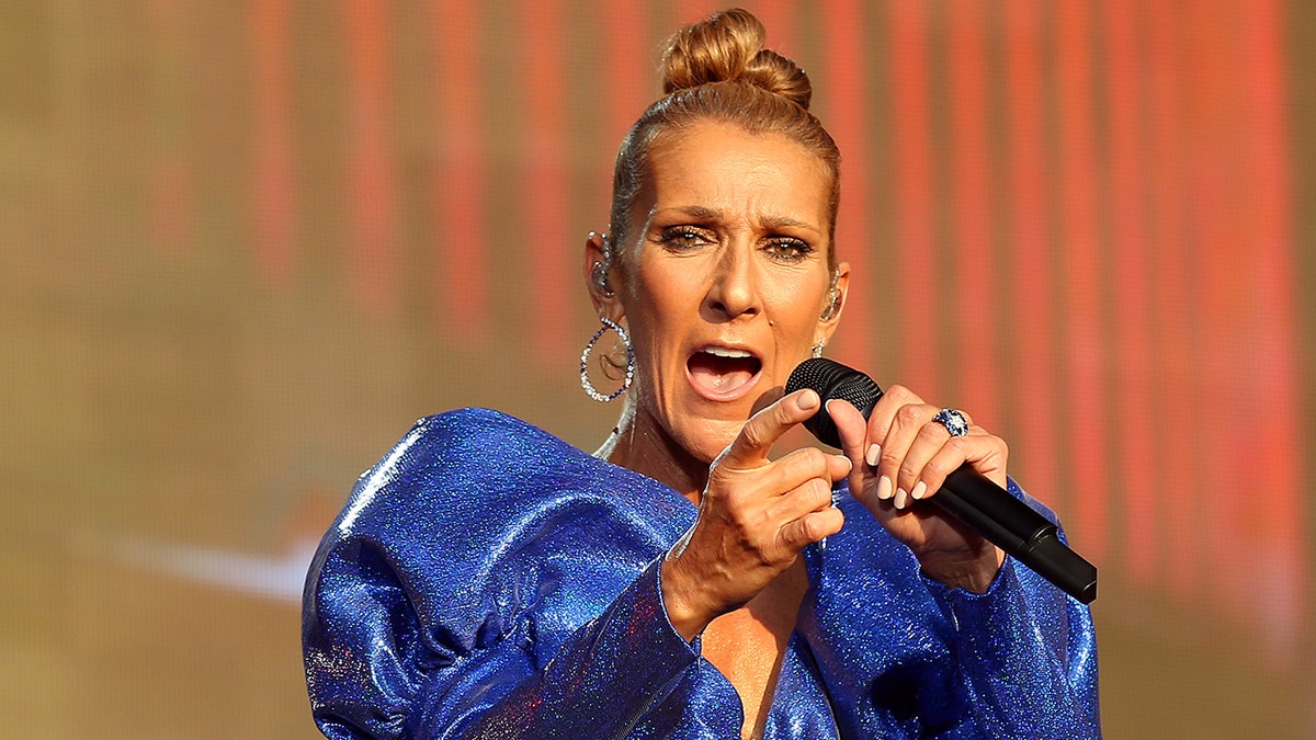 Celine Dion in a blue dress sings into the microphone and points out to the crowd