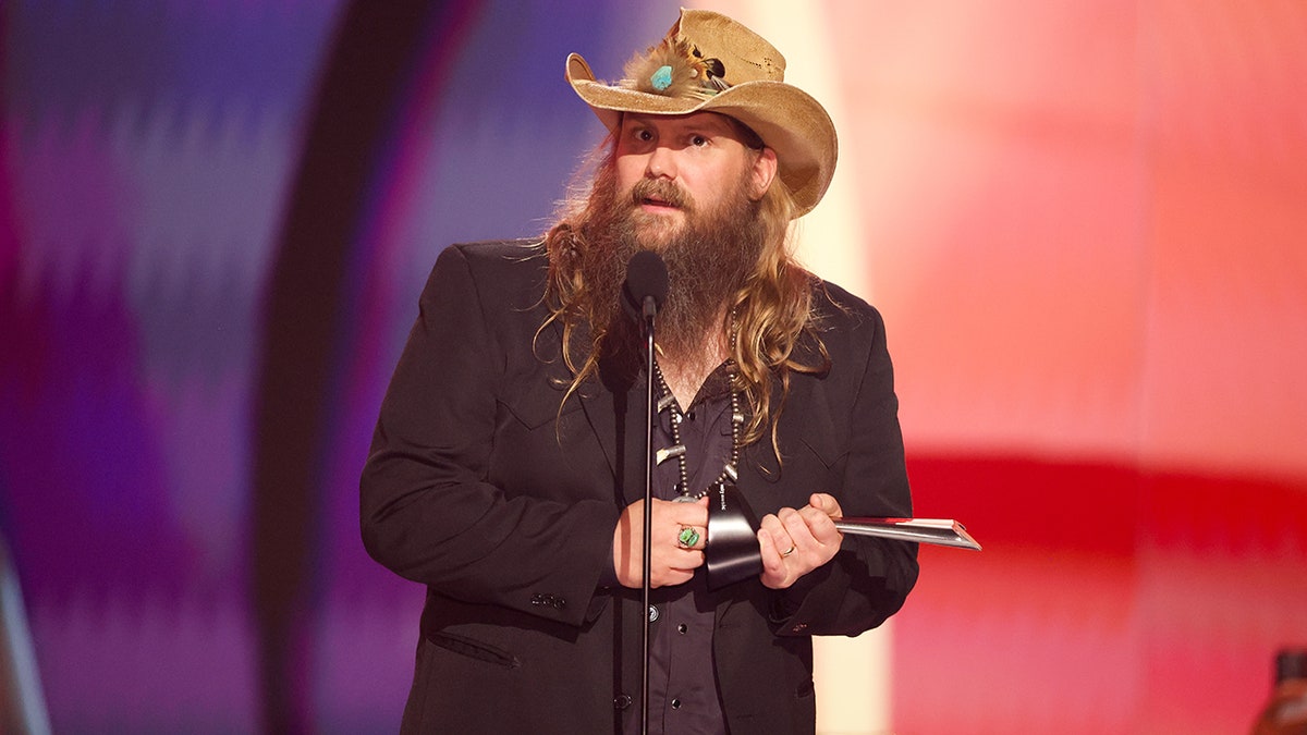 Chris Stapleton de terno e chapéu de cowboy marrom claro no palco do Country Music Awards segura um troféu e fala para a multidão