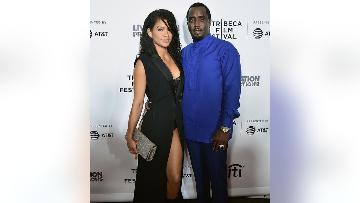 Cassie in a long black dress with a slit stands next to Diddy in a royal blue shirt on the carpet.