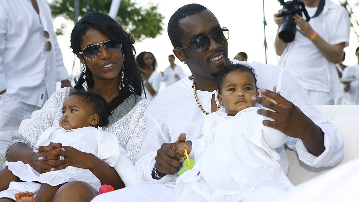 Diddy e Kim Porter, ambos vestidos de branco, segurando suas filhas gêmeas, também de branco