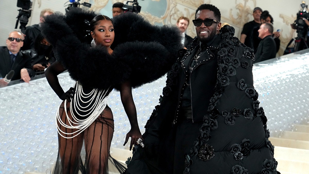 Yung Miami em um vestido de miçangas e um top dramático de penas olha para Diddy no tapete do Met Gala