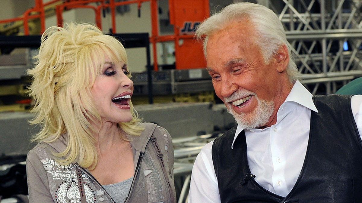 Dolly Parton in a grey sweatshirt laughs with Kenny Rogers in a white shirt and black vest