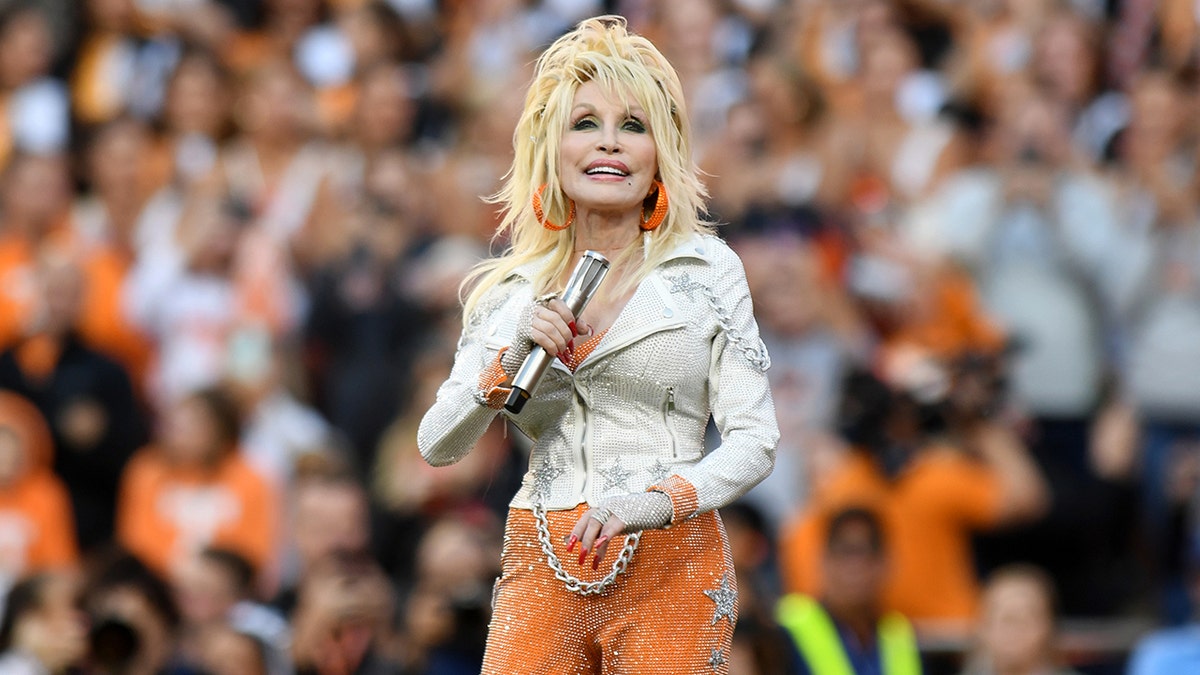 Dolly Parton in a white leather jacket and orange pants holds on to her microphone on the field in Tennessee 