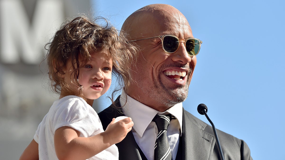 Dwayne Johnson behind the podium in a grey suit and black tie holds daughter Jasmine