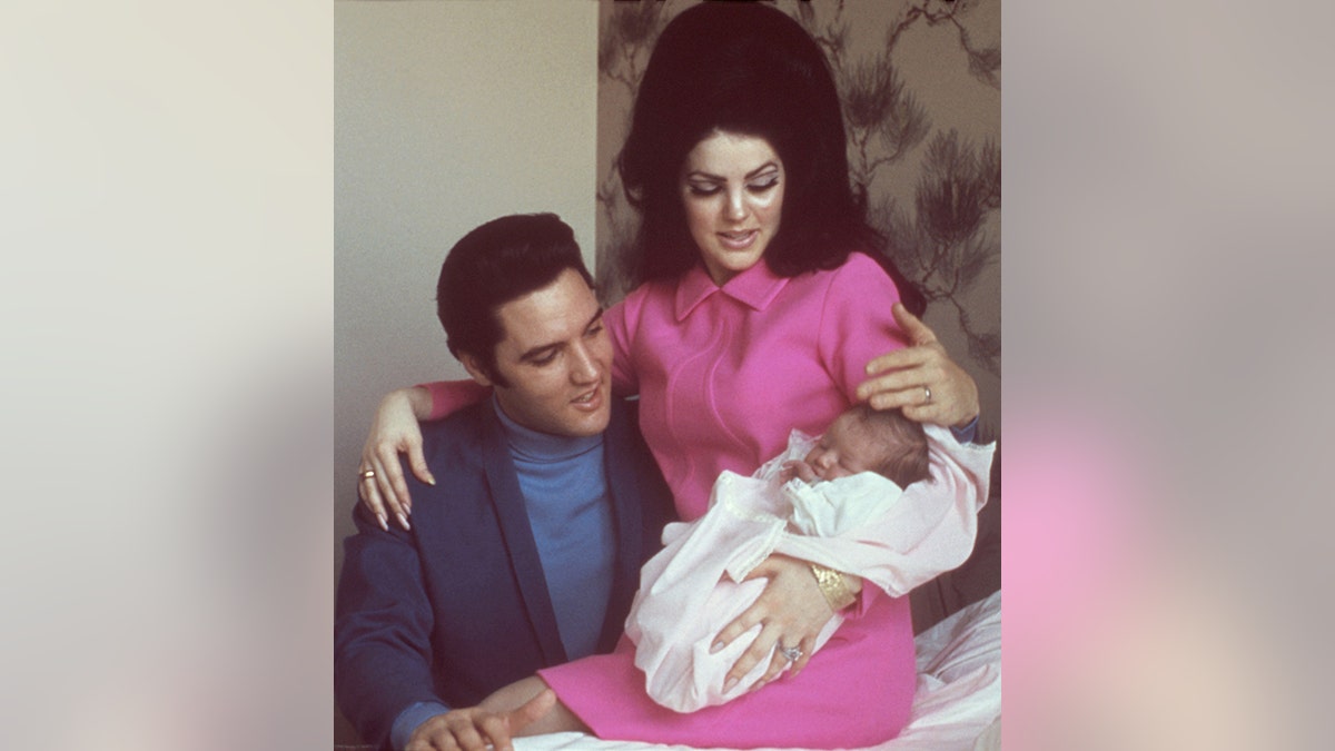 Elvis Presley in a blue shirt and dark blue jacket looks at Lisa Marie Presley as a baby who is being held by Priscilla Presley in pink