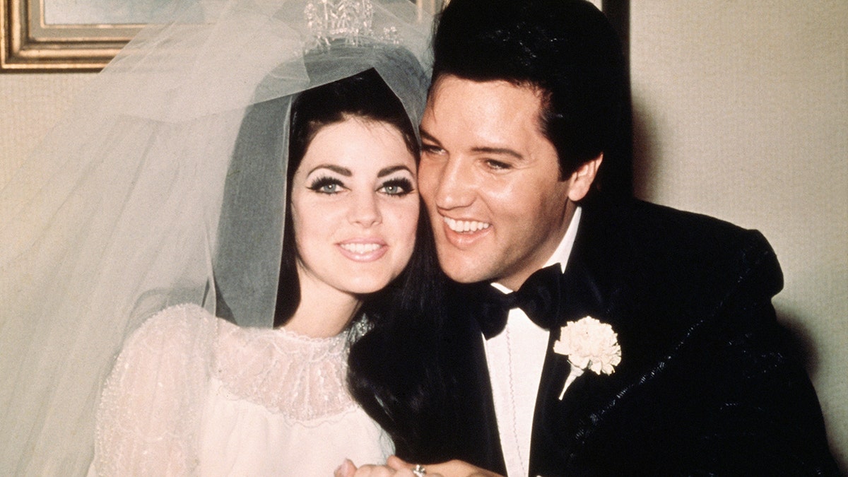 Priscilla Presley em vestido de noiva branco e véu de tule sorri ao lado de Elvis Presley em smoking clássico