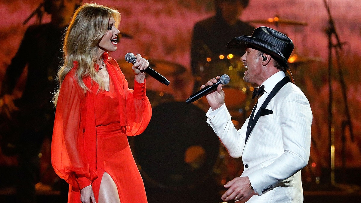 Faith Hill in a long red dress sings to husband Tim McGraw in a white tuxedo and black cowboy hat on stage at the CMA Awards