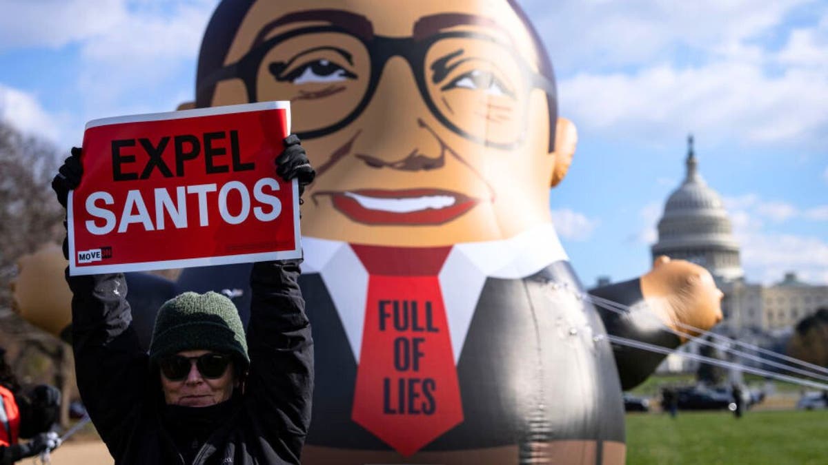 A woman holds a sign in the air reading "expel Santos" while a balloon depicting the Congressman flies in the background in front of the U.S. Capitol