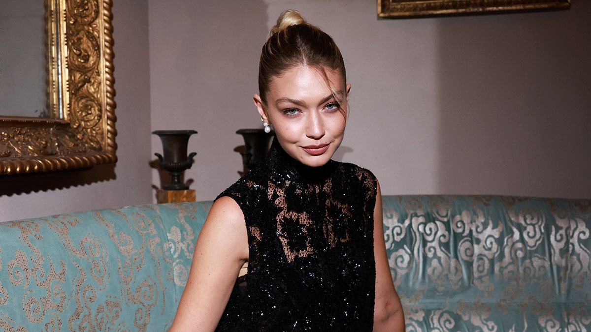 Gigi Hadid in a black lace dress pouts at the camera while sitting on a couch