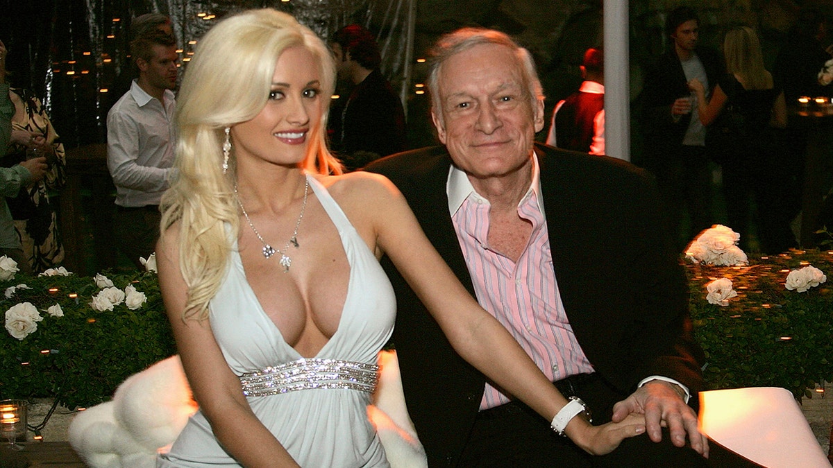Holly Madison in a low cut plunging white dress puts her hand on Hugh Hefners lap as they pose for a photo