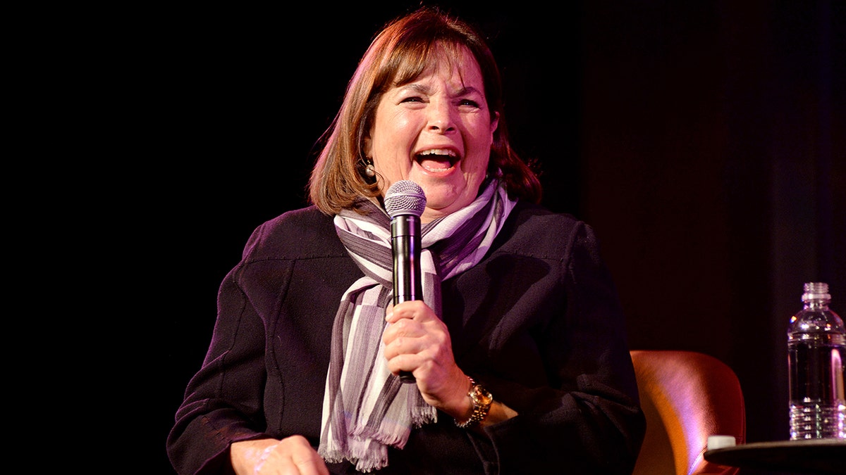 Ina Garten in a dark sweater and striped scarf laughs on stage while holding a microphone