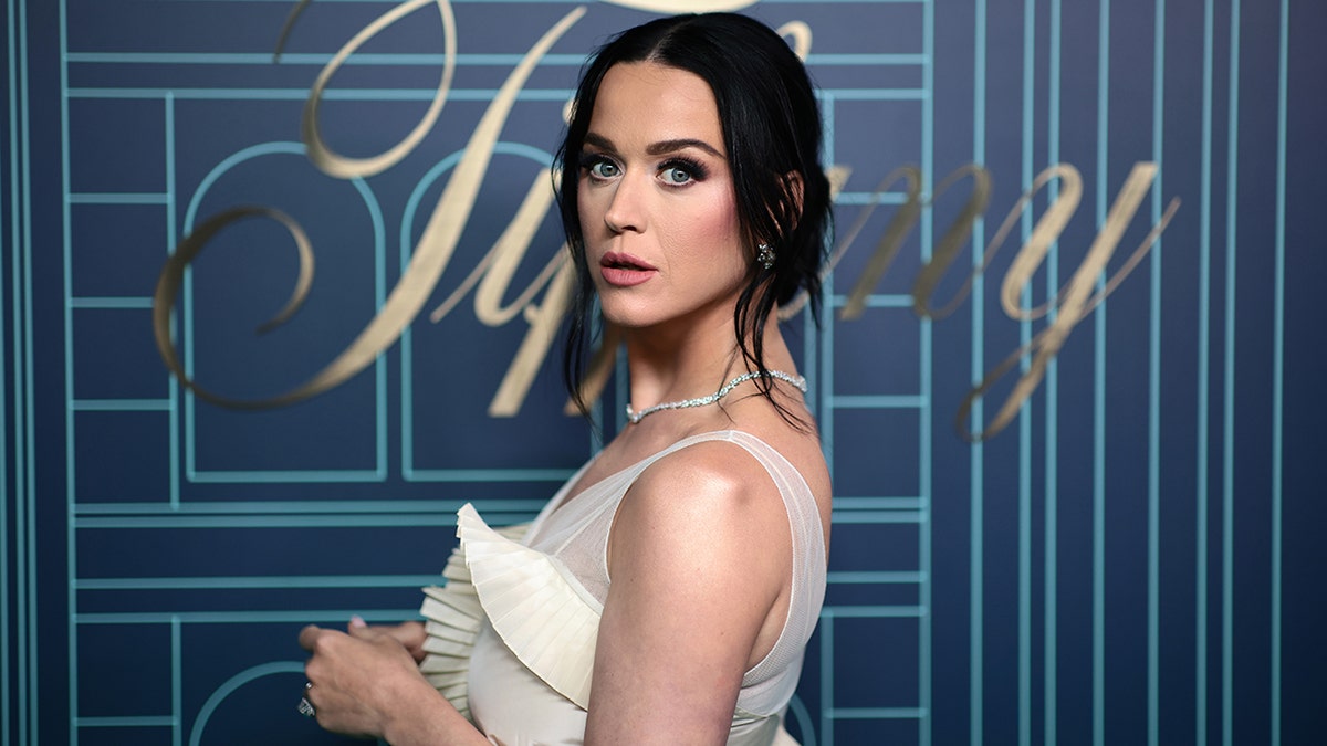 Katy Perry stands on the carpet sideways and looks at the camera in a cream dress