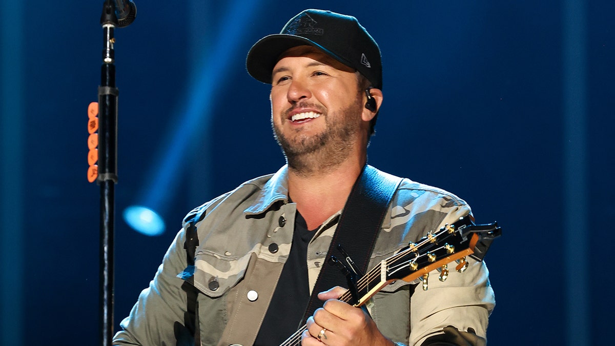 Luke Bryan in a camo jacket and black hat performs on stage with a guitar