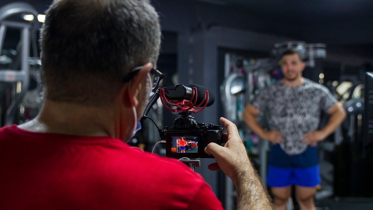Man posing for the camera while vlogger man films at gym