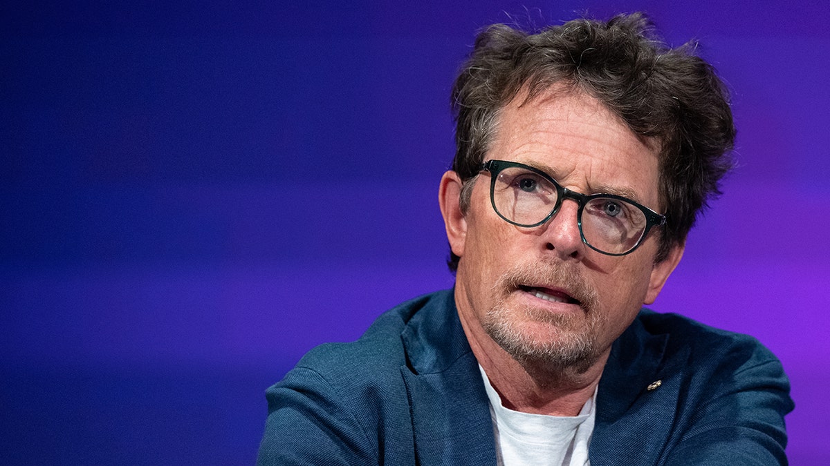 Michael J. Fox in a blue jacket and glasses looks serious on stage
