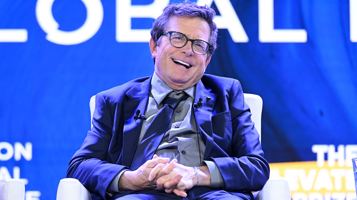 Michael J. Fox sorride sul palco con un abito blu e gli tiene le mani