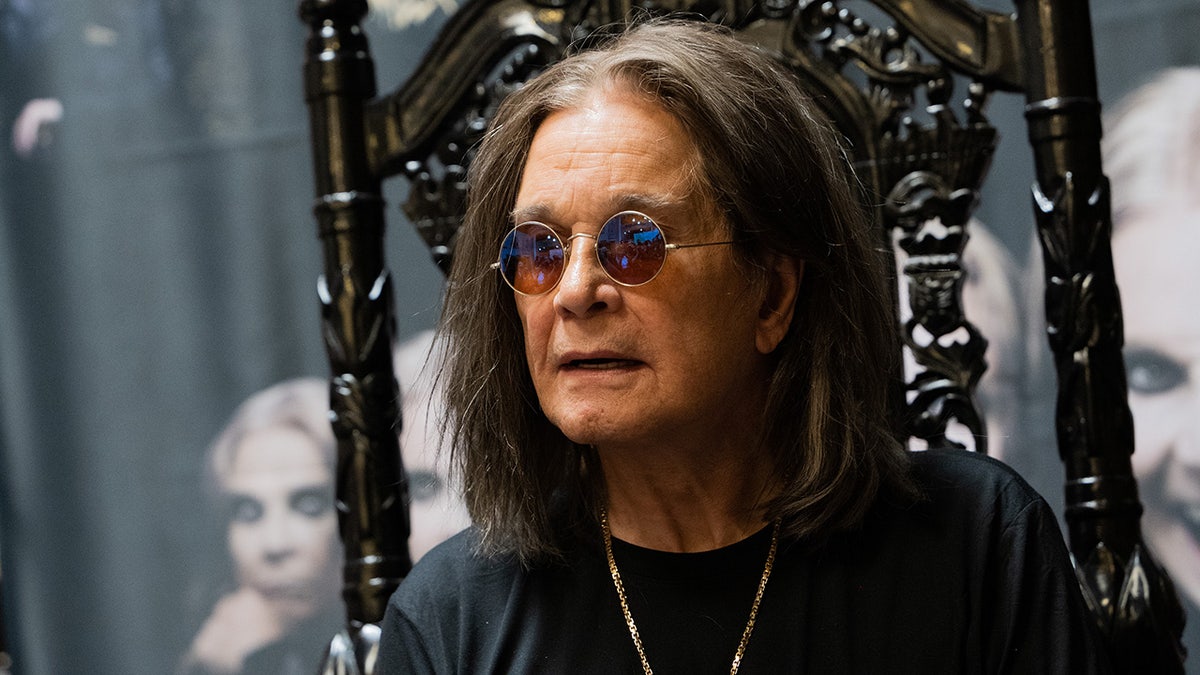 Ozzy Osbourne in black with a gold chain wears circular glasses