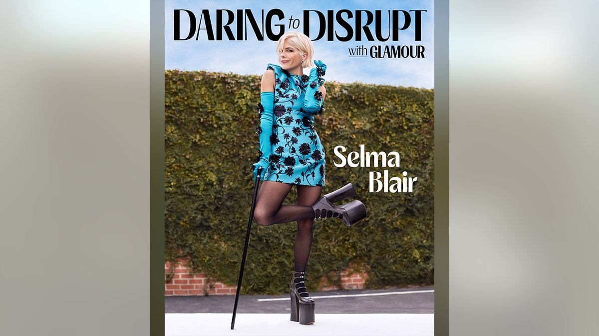 Selma Blair in un abito a fantasia turchese tiene in mano un bastone e alza la gamba destra