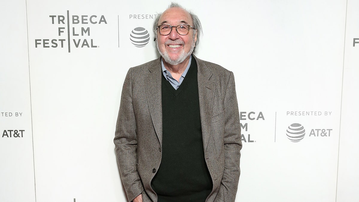James L. Brooks in a grey suit and black sweater smiles with circular glasses on the carpet in New York