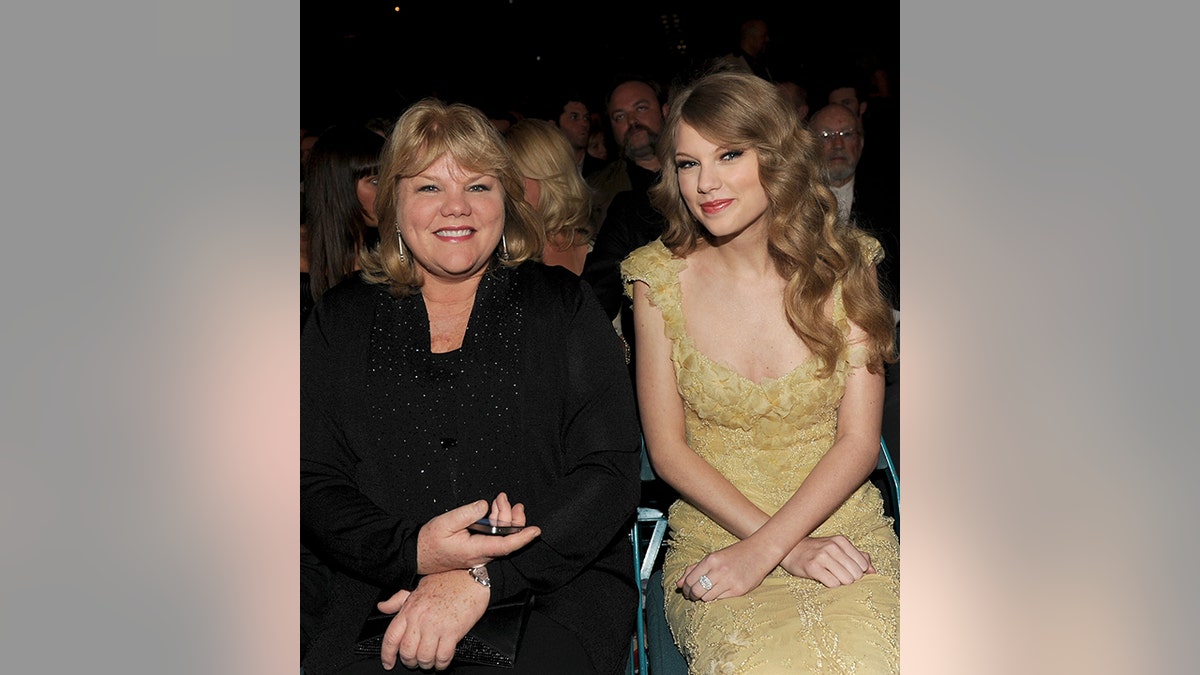 Andrea Swift in all black is seated next to Taylor Swift in a gold dress