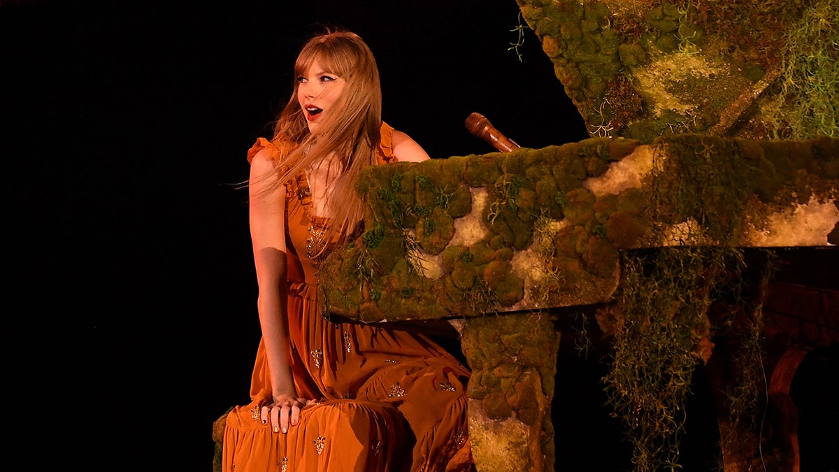 Taylor Swift in an orange dress looks in disbelief towards her crowd while sitting at a piano covered in moss