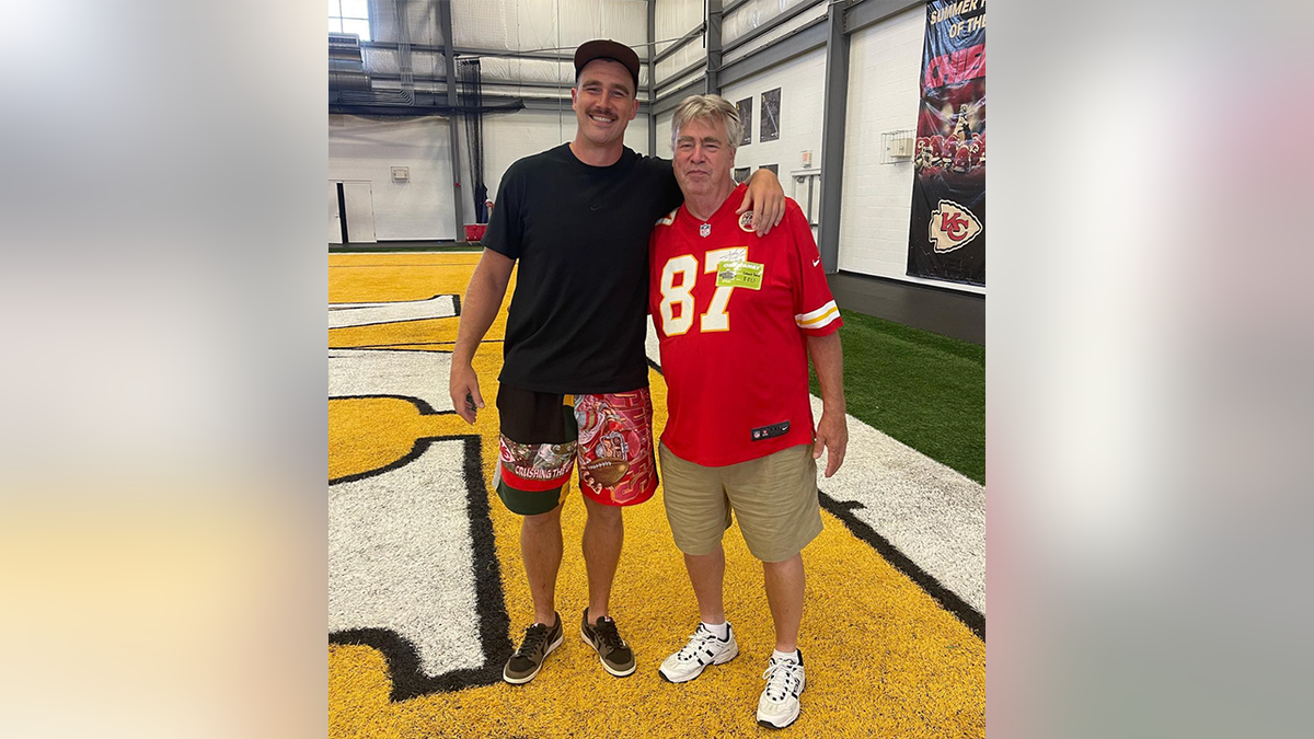 Travis Kelce in a black shirt and Kansas City Chiefs shorts smiles with his father Ed in a Travis Kelce jersey