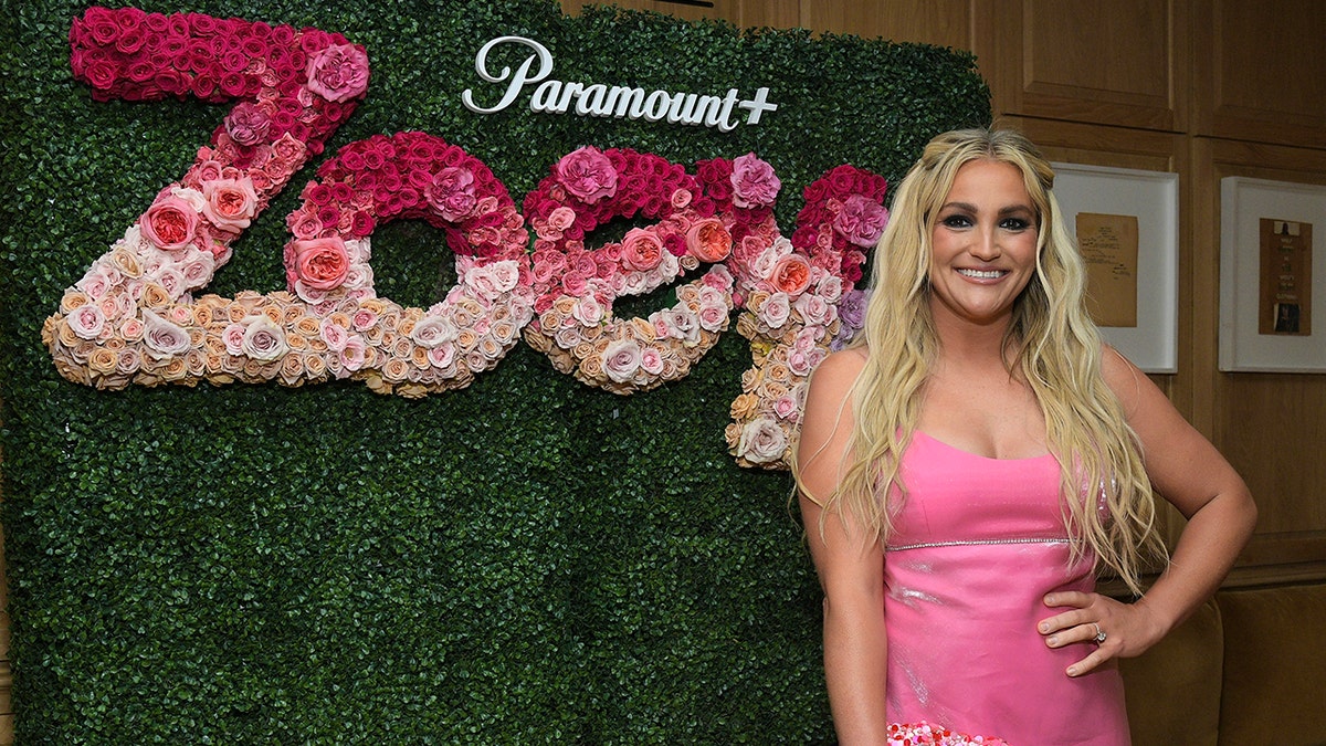 Jamie Lynn Spears in a pink dress puts her hand on her hip standing in front of a green wall with Zoey spelled out in flowers