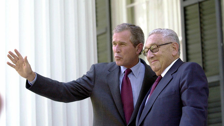 George W. Bush waves to journalists as he stands next to Henry Kissinger