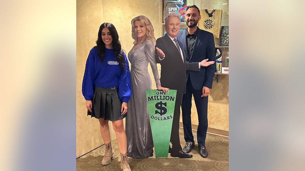 Gishma Tabari in a blue sweater poses with cardboard cutouts of Pat Sajak and Vanna White with her husband in a blue blazer