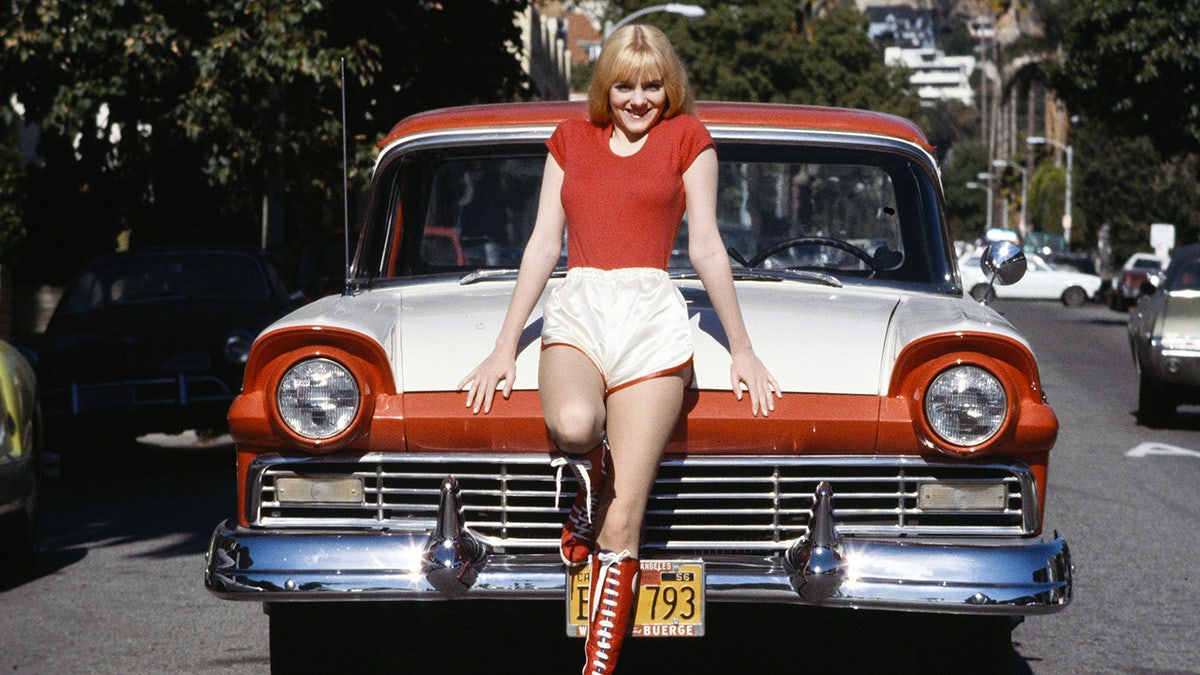 Alison Arngrim in a red shirt and white shorts posing in front of a matching car
