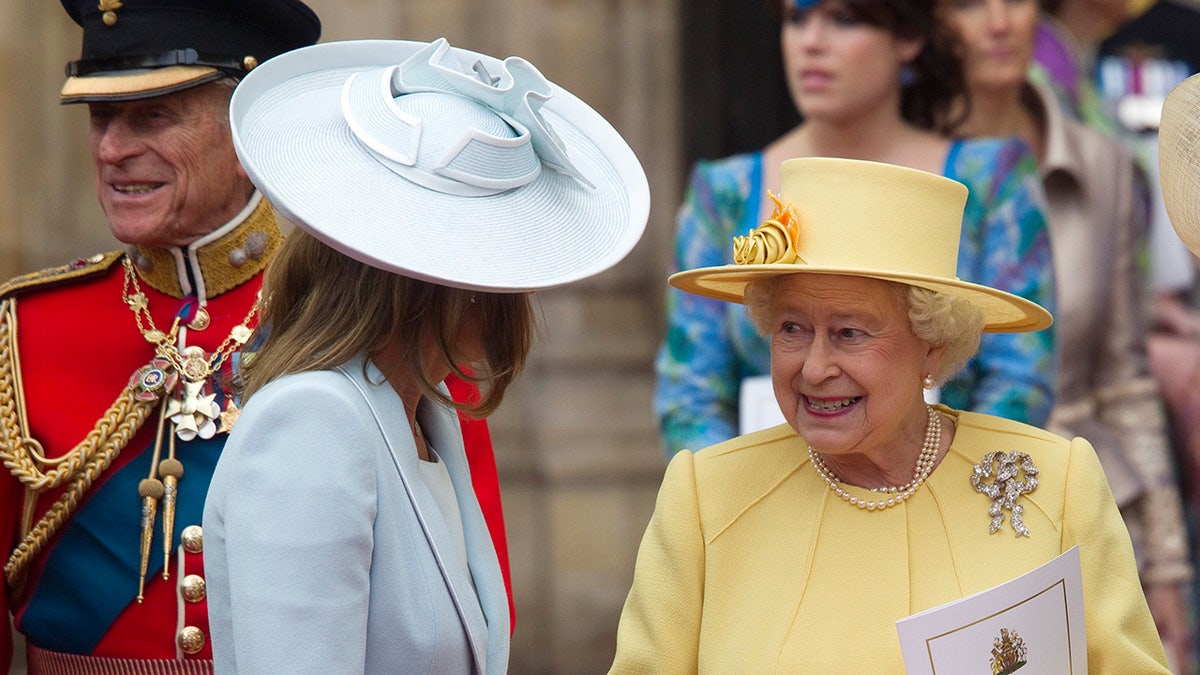 Carole Middleton in a powder blue dress and matching hat speaking to Queen Elizabeth II wearing a yellow dress and matcing hat