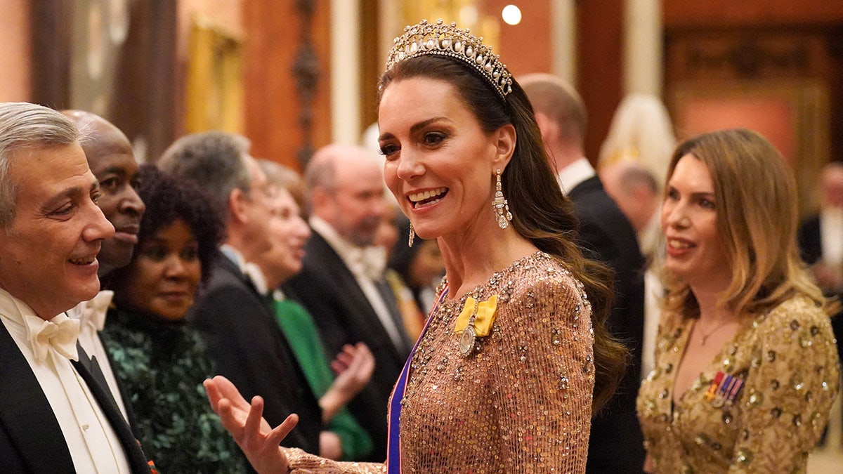 Kate Middleton wearing a sparkling pink dress, a blue and red sash and a lovers knot tiara chatting with people