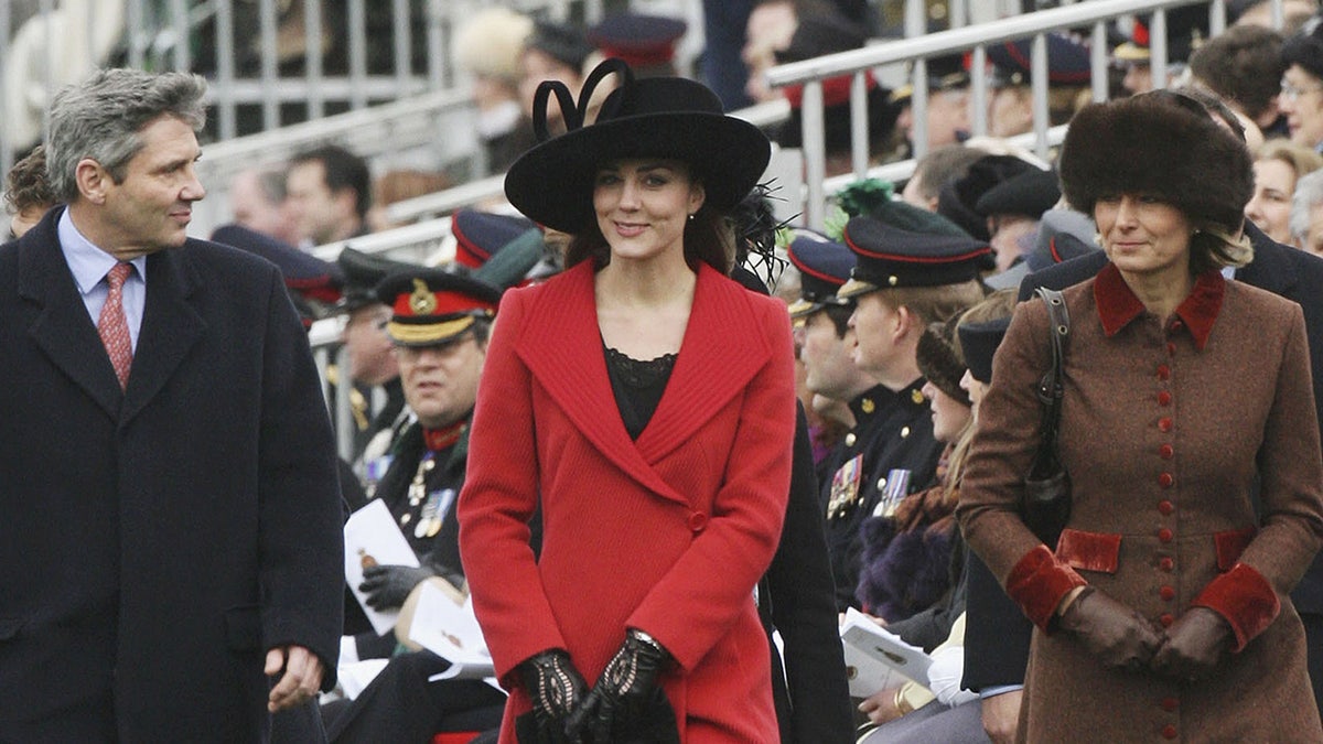 Kate Middleton in a red coat and black hat walking in between her parents