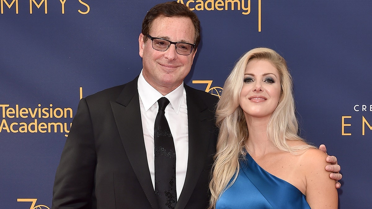 Bob Saget in a black suit and tie next to Kelly Rizzo in a blue one-shoulder satin dress
