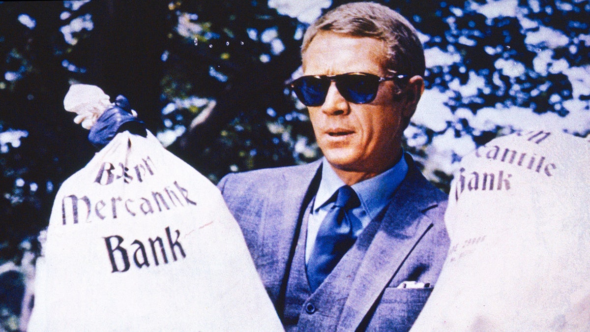 Steve McQueen wearing a blue suit, sunglasses and holding bags of money