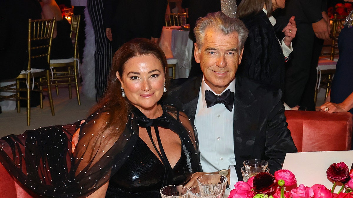 Keely Shaye in a black dress with a mesh shoulder cape sits next to Pierce Brosnan in a traditional tuxedo