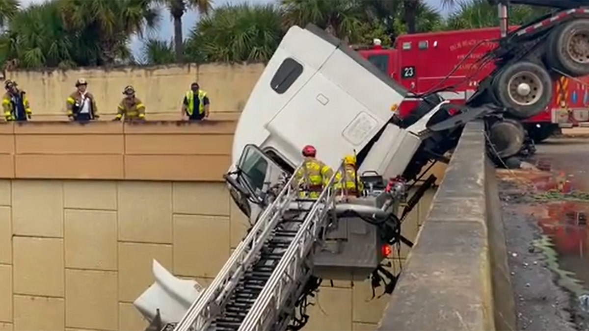 Truck cab dangling over bridge with firefighters on a ladder