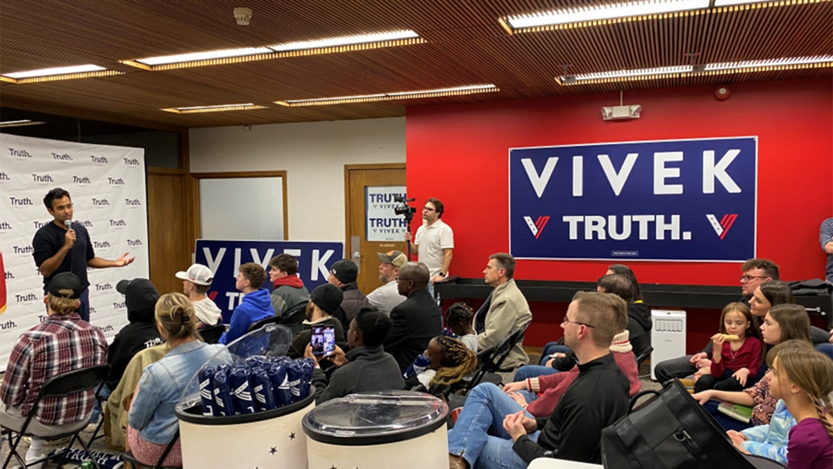 Vivek Ramaswamy speaks to a crowd of supporters in Iowa, holding a microphone surrounded by Vivek Truth campaign posters and backdrop