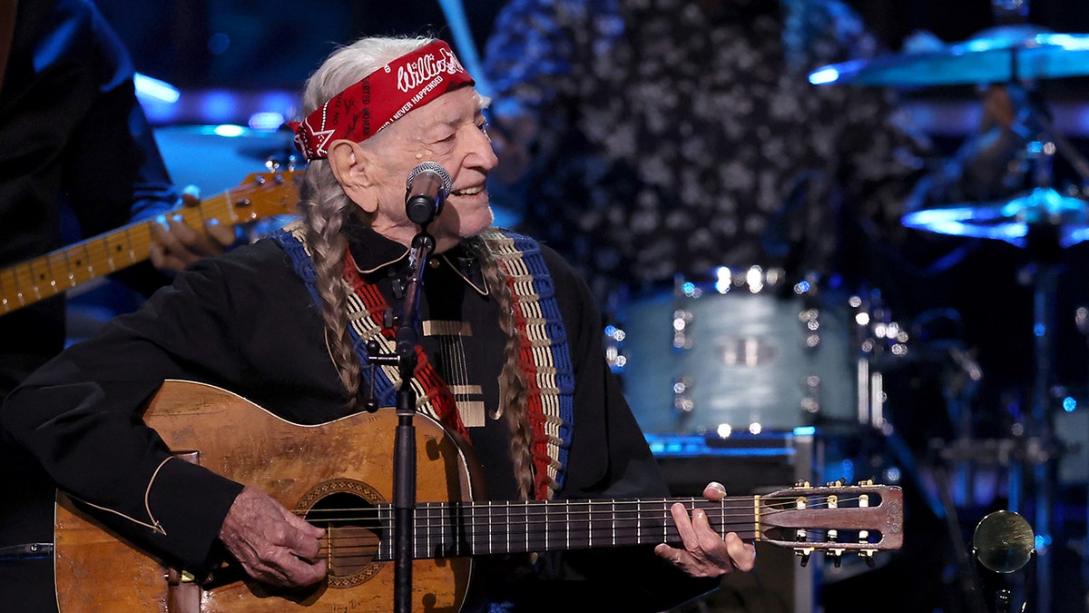 Willie Nelson sitting on stage with a guitar and a microphone