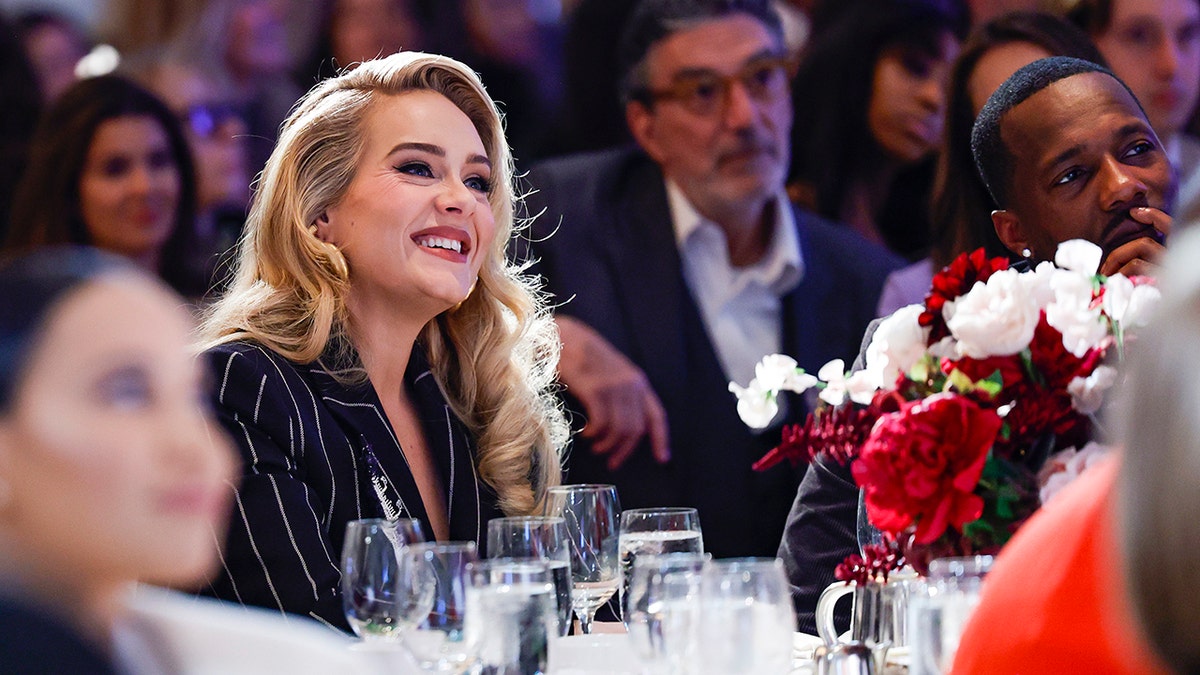 Adele com uma jaqueta listrada está sentada em uma mesa sorrindo ao lado do namorado Rich Paul