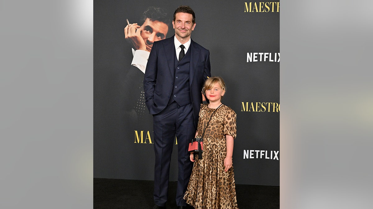 Bradley Cooper em um terno azul-marinho sorri com a filha Lea em um vestido de leopardo no tapete para "Maestro"