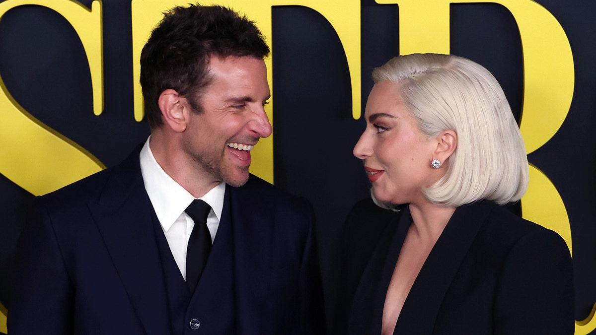 Bradley Cooper sorri e olha com adoração para Lady Gaga em uma roupa preta