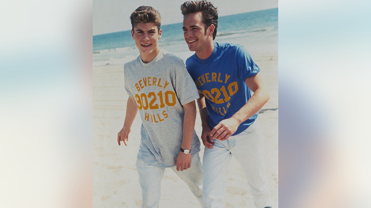 Brian Austin Green com uma camisa cinza Beverly Hills 90210 e Luke Perry com uma camisa azul Beverly Hills 90210 caminham juntos na praia