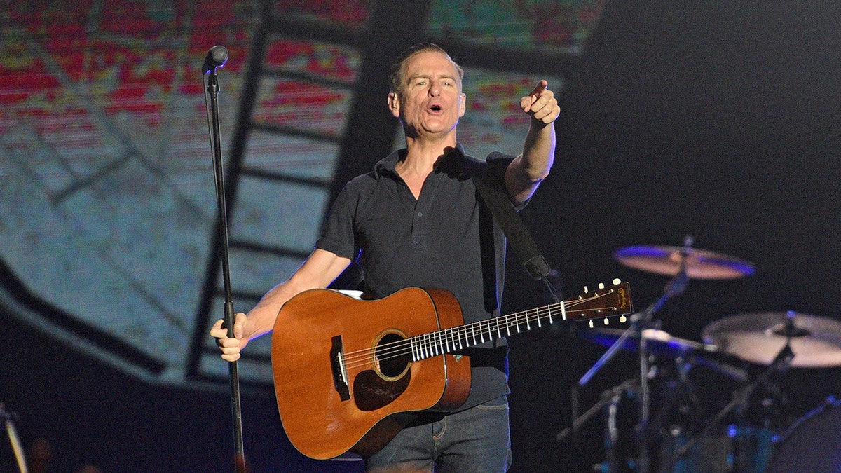 Bryan Adams in a black shirt and dark pants points out to the audience while playing on stage