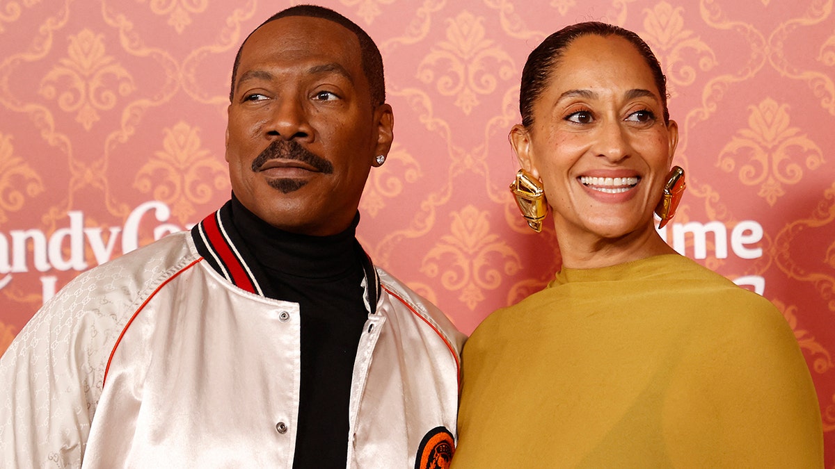 Eddie Murphy in a white jacket looks to his right on the red carpet as he poses with Tracee Ellis Ross in a mustard yellow dress