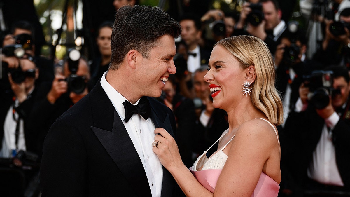 Colin Jost in a black tuxedo laughs on the carpet with wife Scarlett Johansson in a pink dress in Cannes