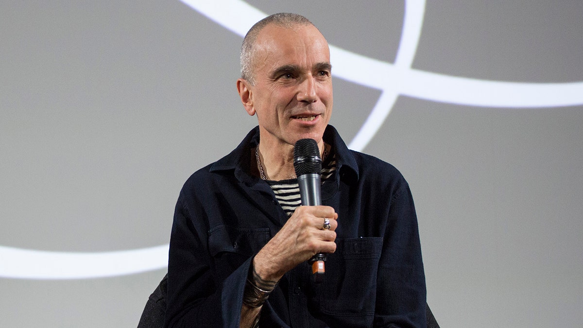 Daniel Day Lewis speaks on stage and holds a microphone wearing a black sweater