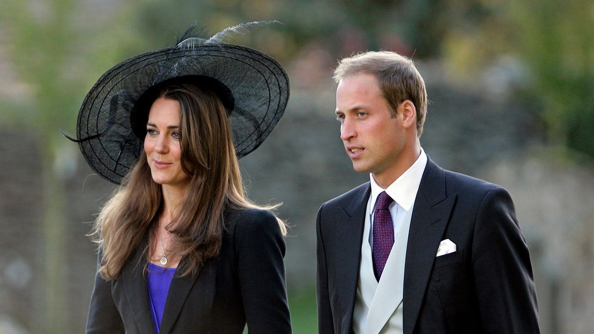 Kate Middleton wearing a black blazer and a bright blue blouse with a black oversized hat standing next to Prince William in a suit and tie